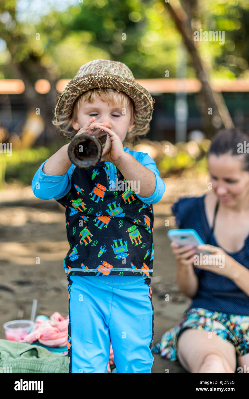 A portrait of a toddler wearing a hat drinking from a brown beer bottle while his mother is intently watching the smartphone Stock Photo