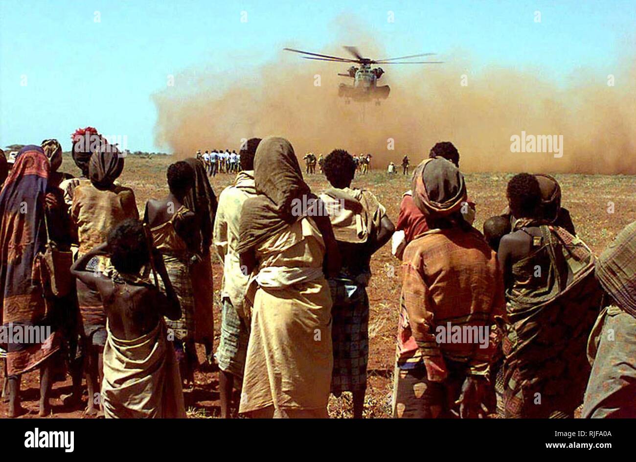 The camera shoots past some Somalis from the village of Maleel as they watch a US Marine CH-53 Sea Stallion deliver a sling load of wheat donated by the people of Australia.  Several Somali men and Australian Army Soldiers wait near the loading zone where the helicopter rotor wash is kicking up large amounts of red dirt.  This mission is in direct support of Operation Restore Hope. Stock Photo
