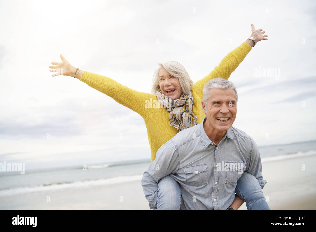 Modern vibrant senior couple piggy back riding on beach Stock Photo