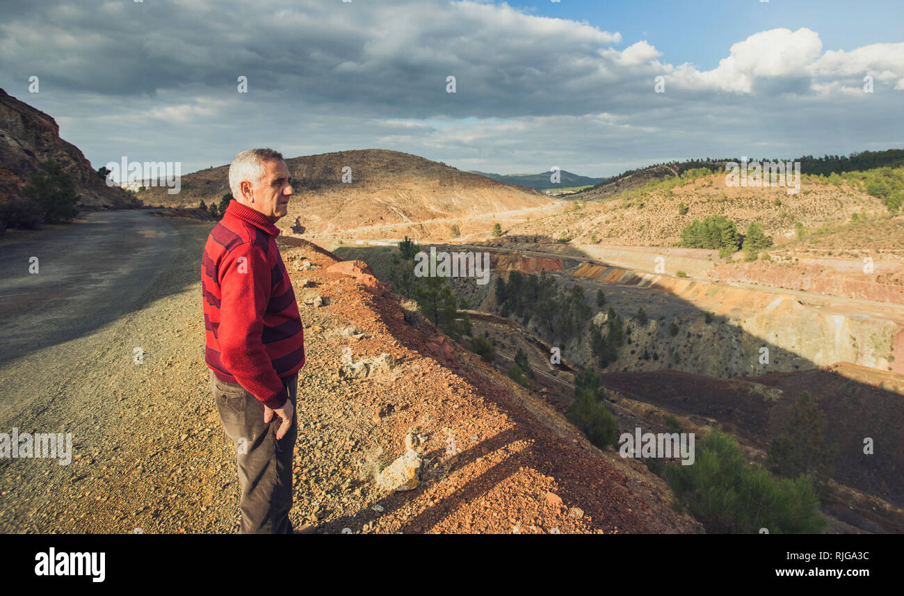 Side portrait of an elderly man with white hair and red striped blouse, with the Zaranda mines and sky cloudy in the background, Spain Stock Photo