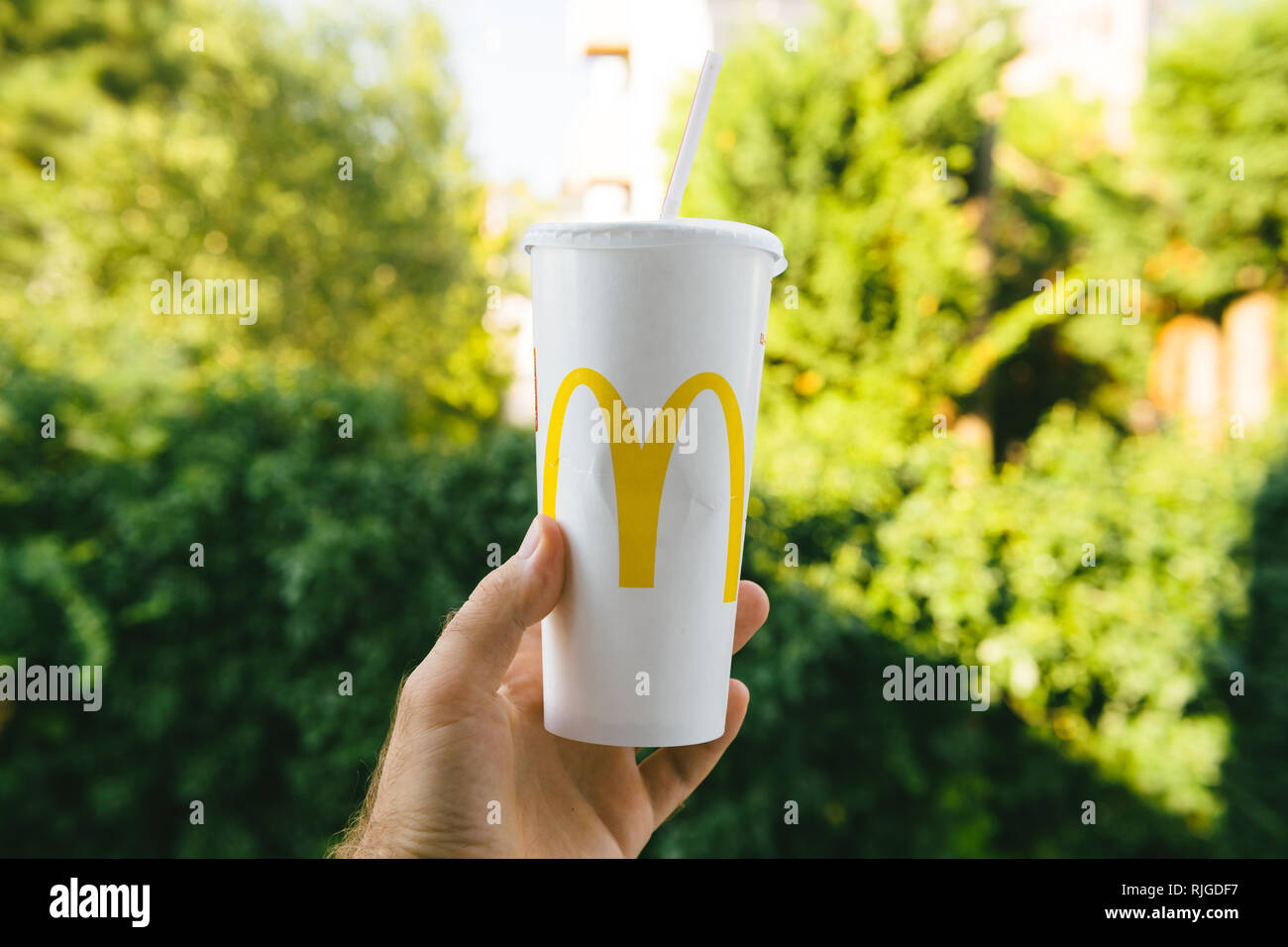 PARIS, FRANCE - JUL 27, 2018: Man holding outdoor against green background a cup of McDonald's Cola with plastic straw with yellow logo near one of the world's largest fast food restaurants Stock Photo
