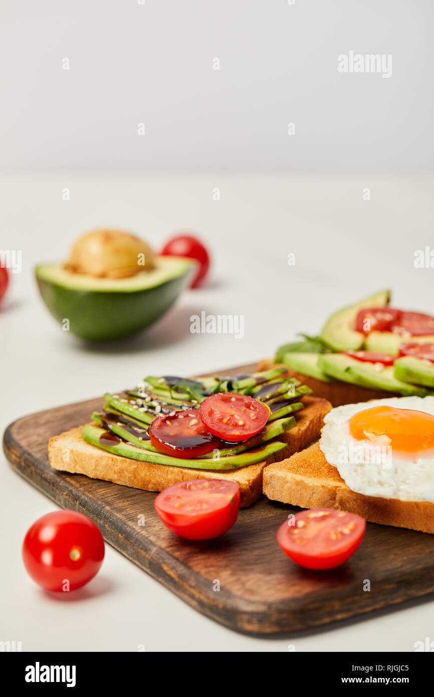 selective focus of wooden cutting board with toasts, scrambled egg, cherry tomatoes and avocado on grey background Stock Photo