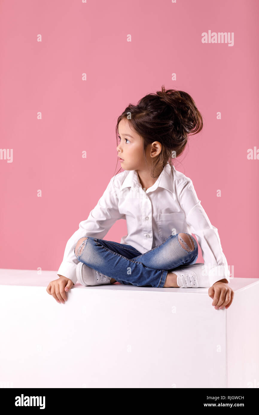 Cute calm little child girl in white shirt with hairstyle sits in lotus position and looks away on pink background. Stock Photo