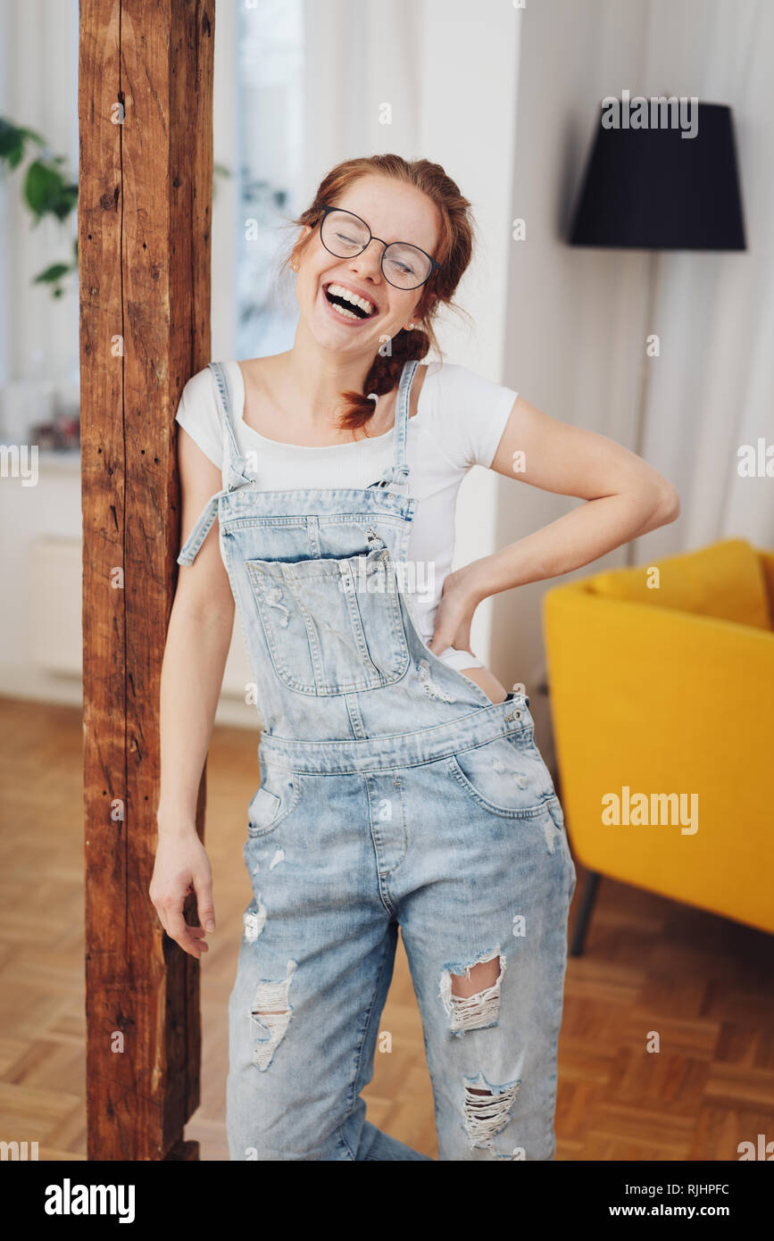 Cute young red-haired girl with pigtails and glasses, standing indoors in  denim overalls, leaning on wooden pole and laughing Stock Photo - Alamy