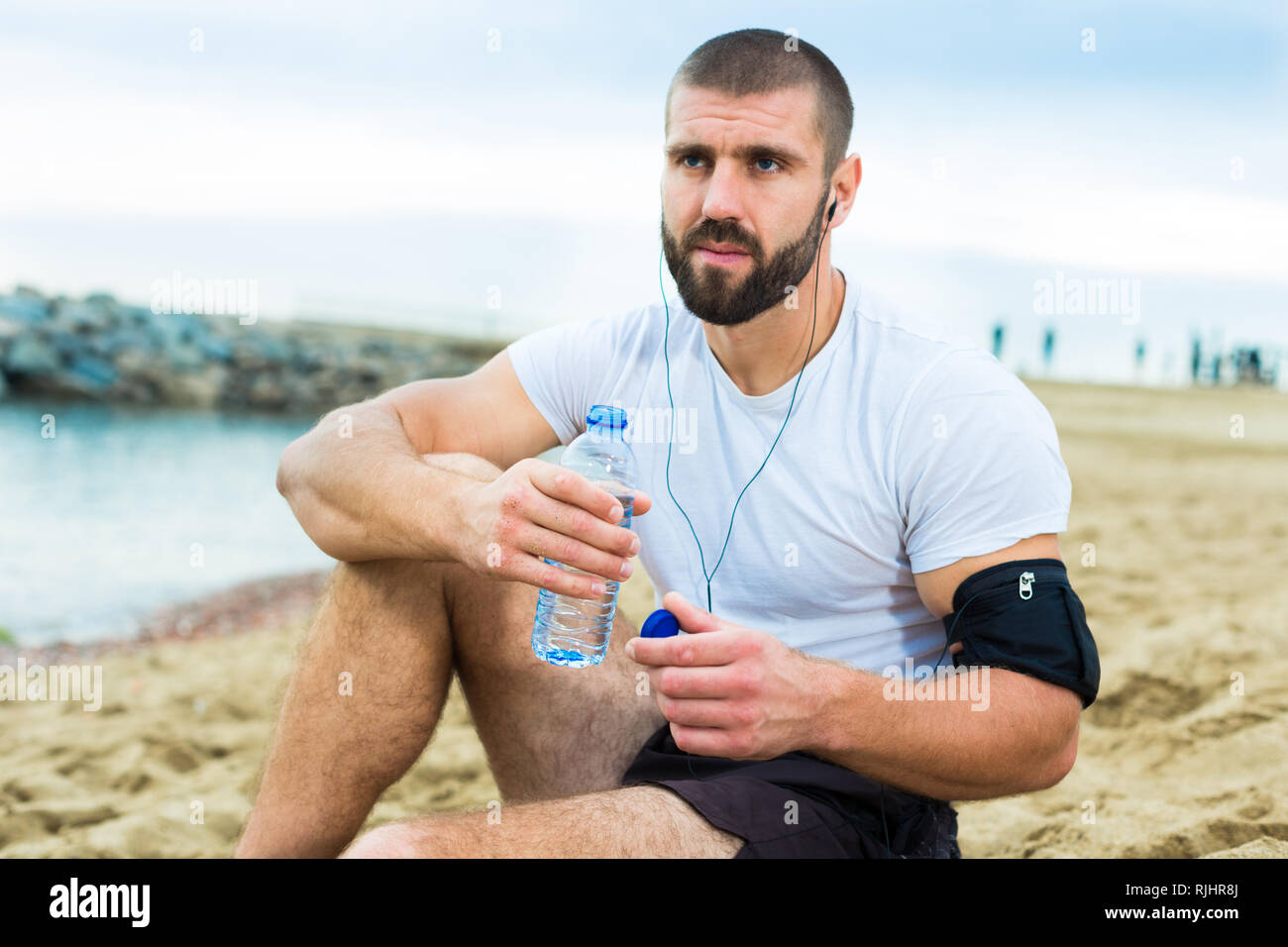 Man in sportswear sits on the sand near the sea shore after an active ...