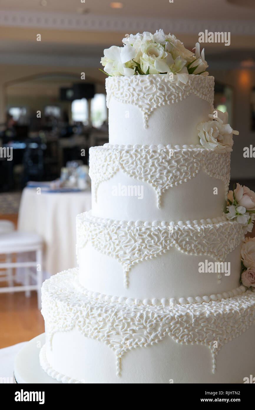 white wedding cake with white flowers and fancy designs with a reception hall in the background Stock Photo