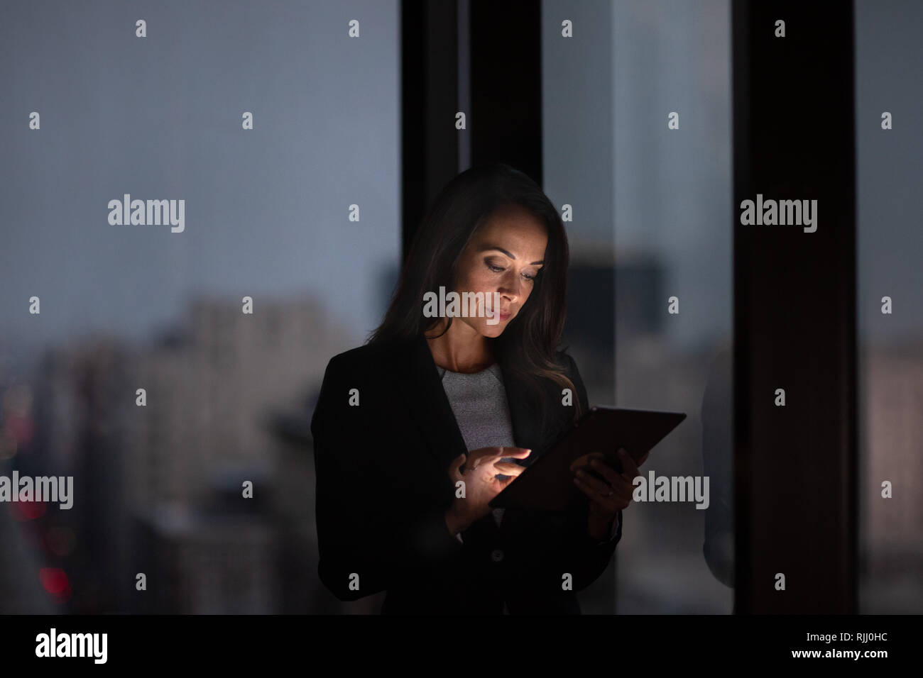 Businesswoman working late using digital tablet with city skyline in background Stock Photo