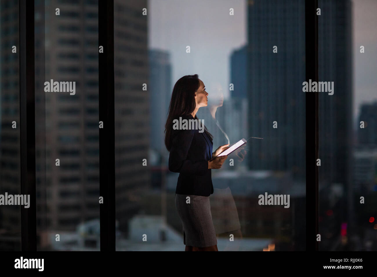 Businesswoman working late using digital tablet with city skyline in background Stock Photo
