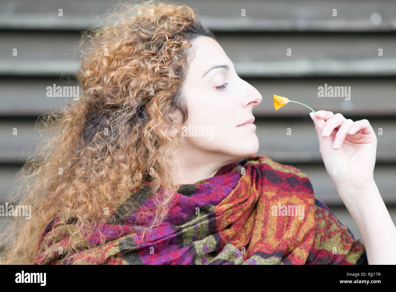 profile portrait of sultry beautiful woman with long red windswept curly hair and brown eyes sniffing a fragrant buttercup flower Stock Photo