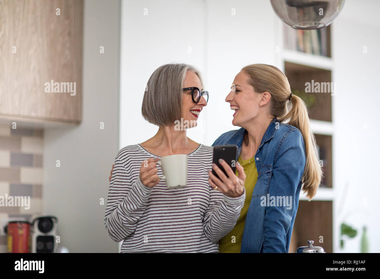 Mature adult woman with grown up daughter looking at smartphone in kitchen Stock Photo