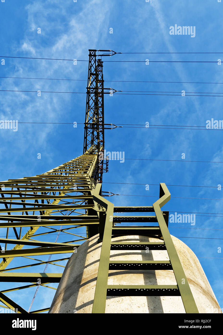 View from below up to a high voltage pylon, which stands on a concrete foundation, a ladder leads up. Above it a blue sky with light clouds of fog. Stock Photo