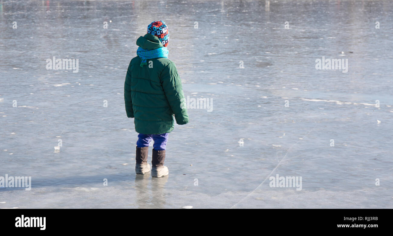 A child standing on a frozen pond bundled up in a winter coat, hat, boots and scarf. Stock Photo