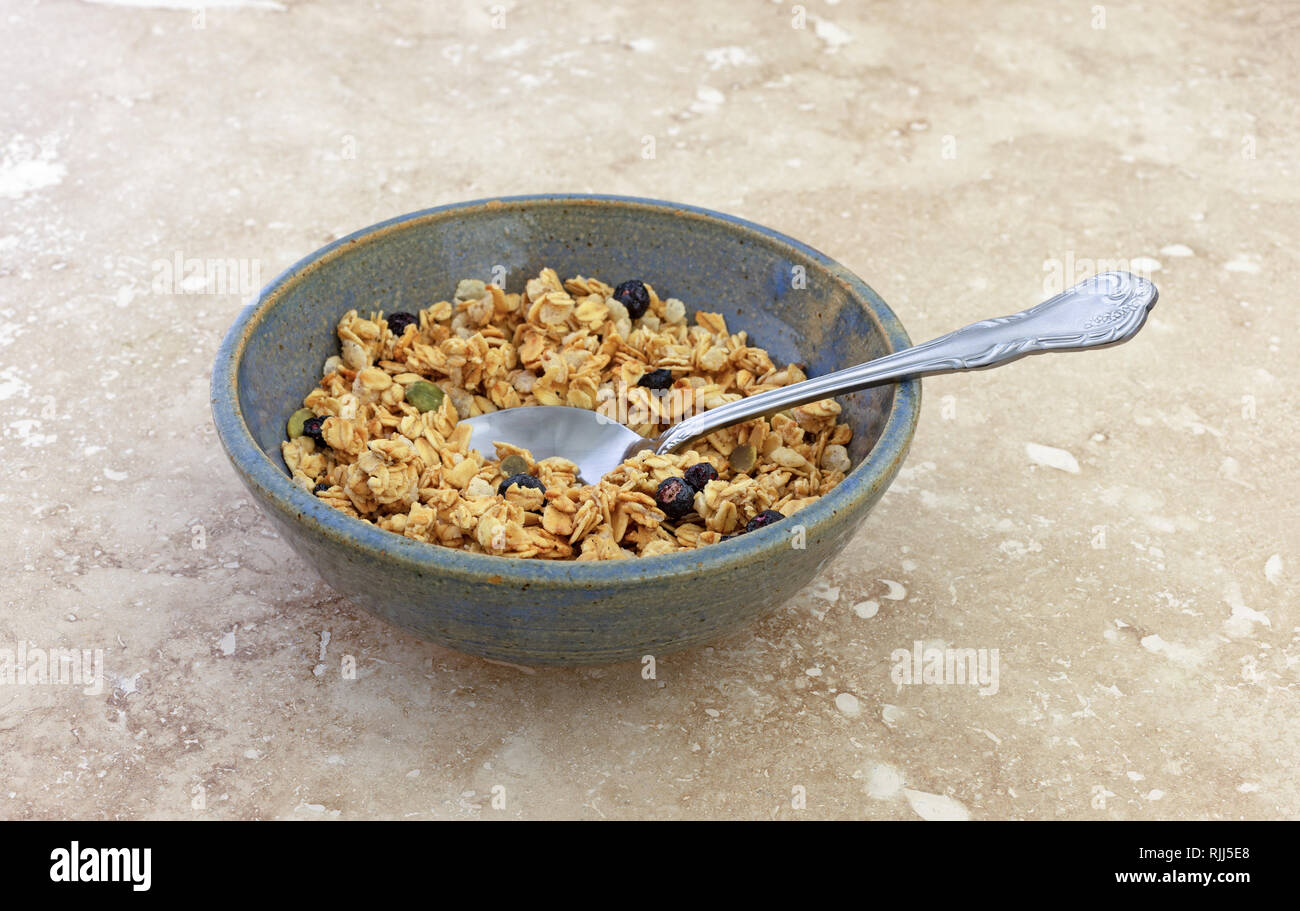 Side view of a bowl of dry organic breakfast cereal with dried blueberries and pumpkin seeds plus a spoon in the food on a marble table. Stock Photo