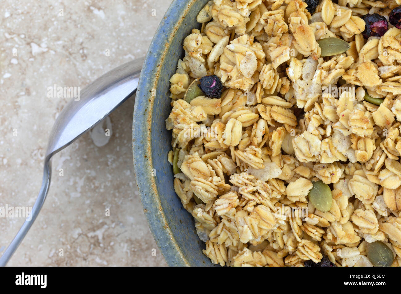 Top close view of a bowl of dry organic breakfast cereal with dried blueberries and pumpkin seeds plus a spoon in the food on a marble table. Stock Photo