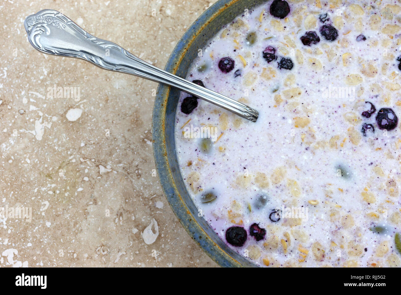 Top close view of organic breakfast cereal with milk dried blueberries and pumpkin seeds plus a spoon in the food on a marble table. Stock Photo