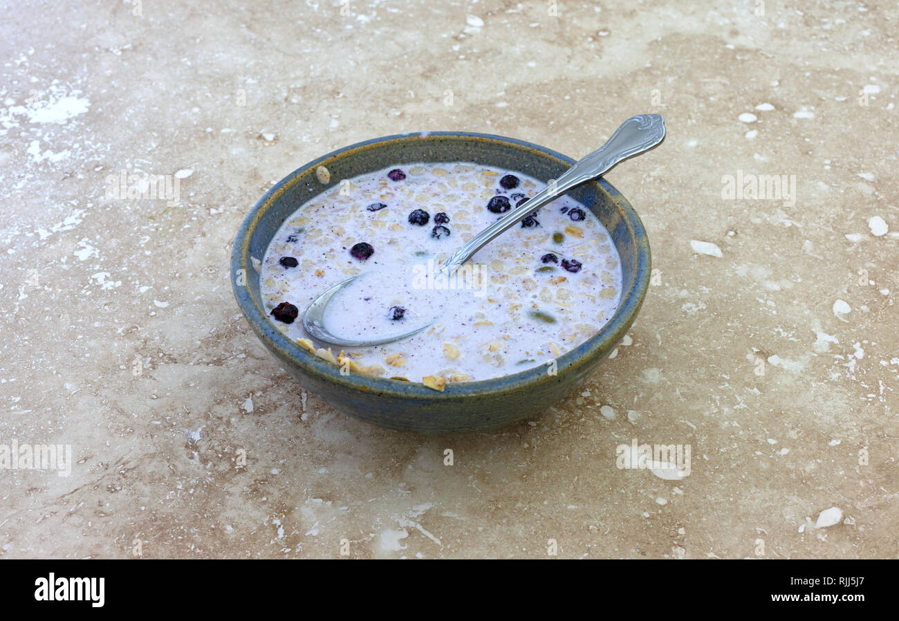 Side view of organic breakfast cereal with milk dried blueberries and pumpkin seeds plus a spoon in the food on a marble counter top. Stock Photo