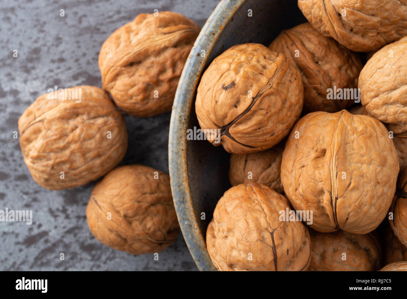 Overhead close view of an old stoneware bowl filled with walnuts plus three nuts to the side on a gray counter top. Stock Photo