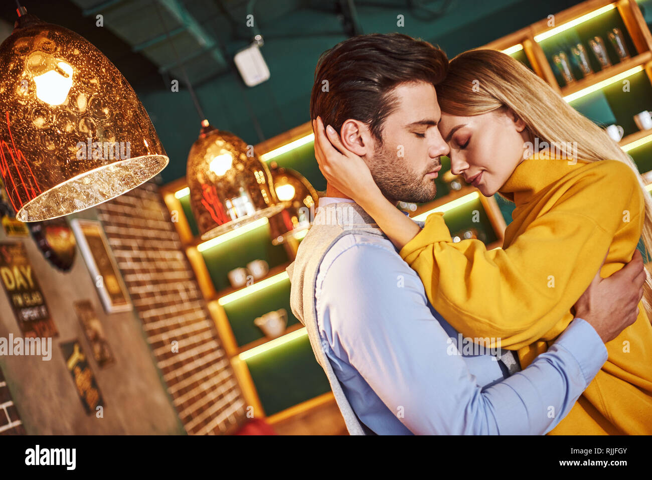 Real love. Young man holding pretty blonde woman in his hands. Close-up of young woman huggs her boyfriend in restaurant. Blonde young woman in mustar Stock Photo