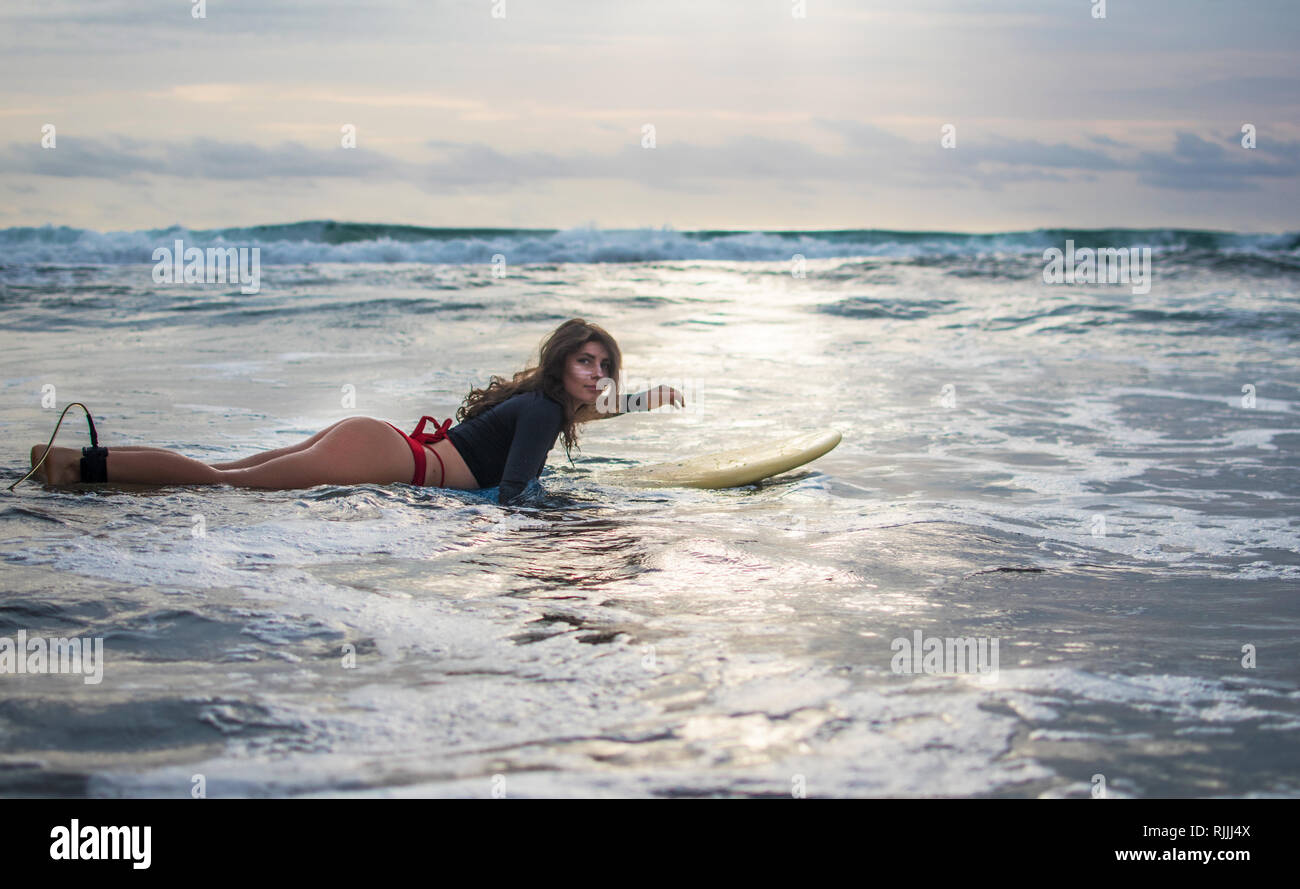 Joyful woman in have fun before surfing. Surfer girl with white lines mask on her pretty face hold surf board, look at the beach. People in sport Stock Photo