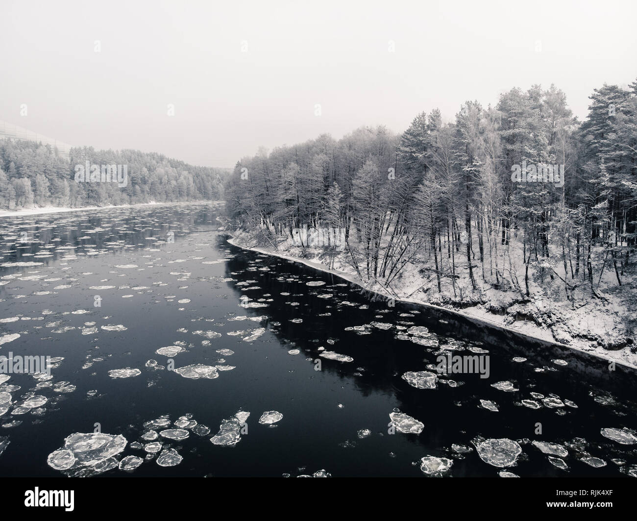 View to the snowy shore from the middle of freezing river with reflections of the forest in water Stock Photo