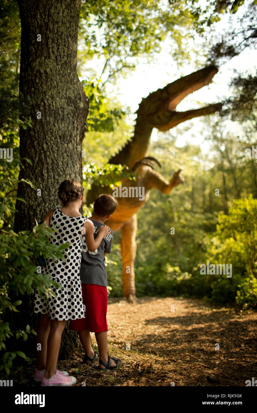 Young brother and sister hiding from a statue of an enormous dinosaur. Stock Photo