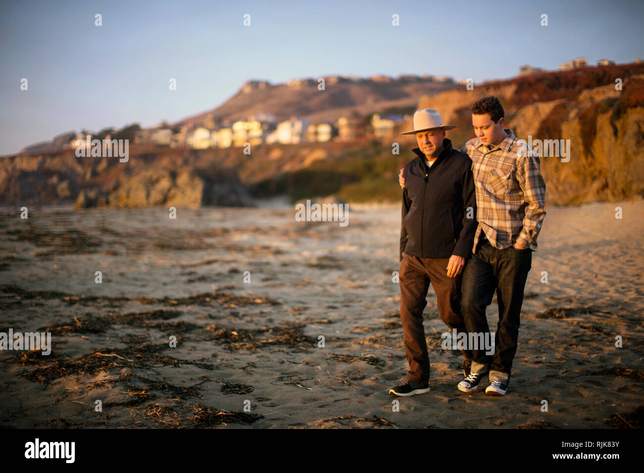 Teenage son puts a comforting arm around his middle-aged father's shoulders as they walk together on the beach at sunset. Stock Photo