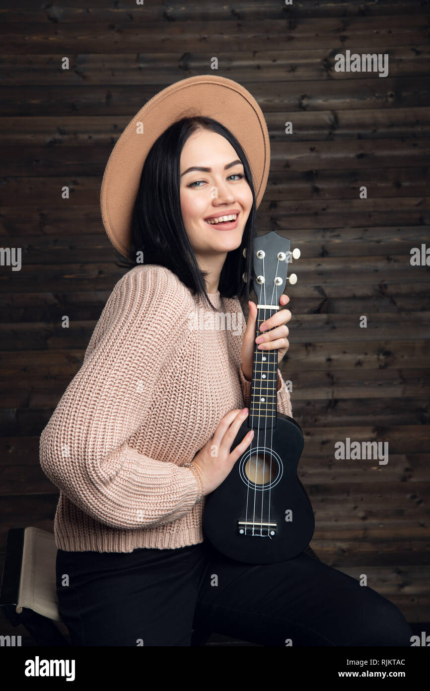 Attractive emotional young Caucasian woman in sweater and hat playing ukulele. Beautiful woman