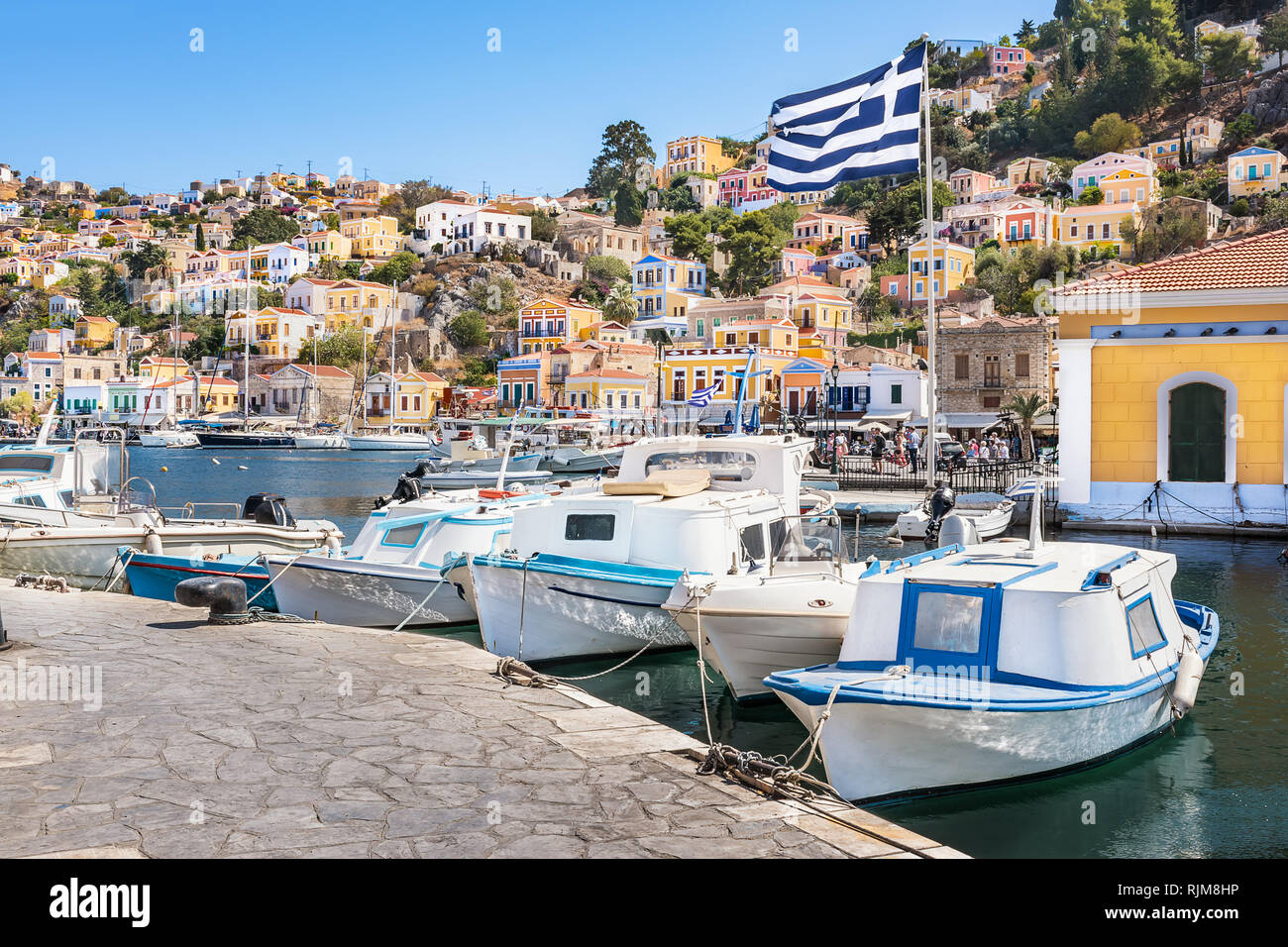 Greek flag, boats and colorful neoclassical houses in harbor town of Symi (Symi Island, Greece) Stock Photo
