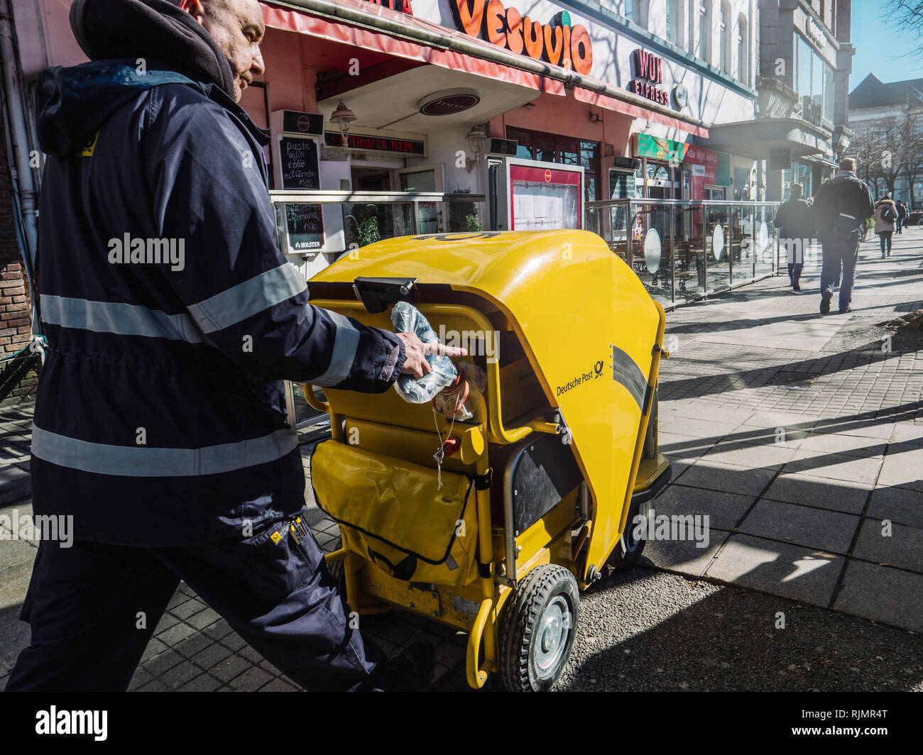 HAMBURG, GERMANY - MAR 20, 2018: Man in uniform pushing Deutsche Bank DHL German post yellow carriage with mail walking down street Stock Photo
