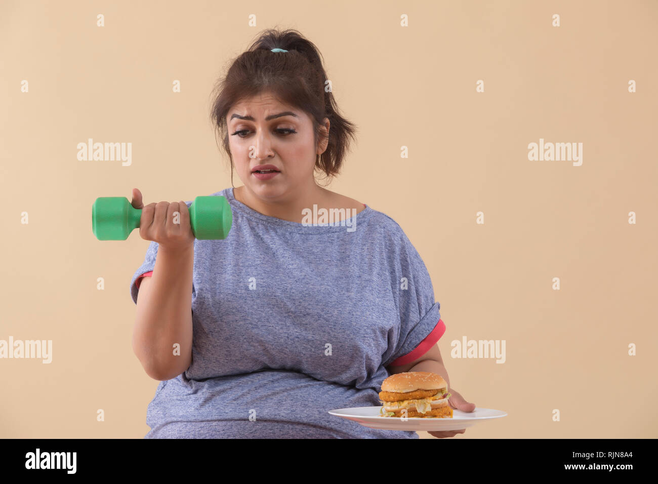 Sad Overweight Woman holding a burger and dumbbell Stock Photo