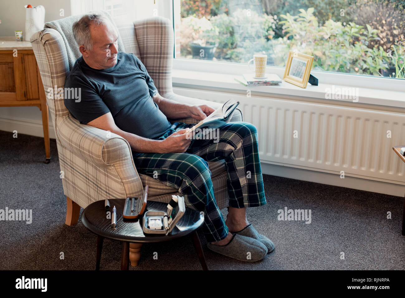 Senior diabetic man is relaxing in the living room of his home with a book. His blood glucose testing kit is on the table next to him. Stock Photo