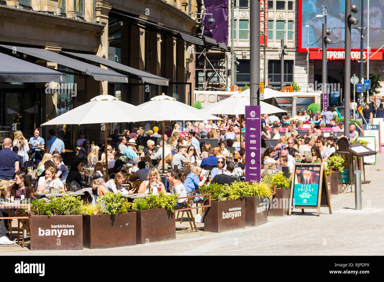 A summer day in Manchester city centre with many people sat eating and drinking outside Banyan Bar and other restaurants and bars of The Triangle, for Stock Photo