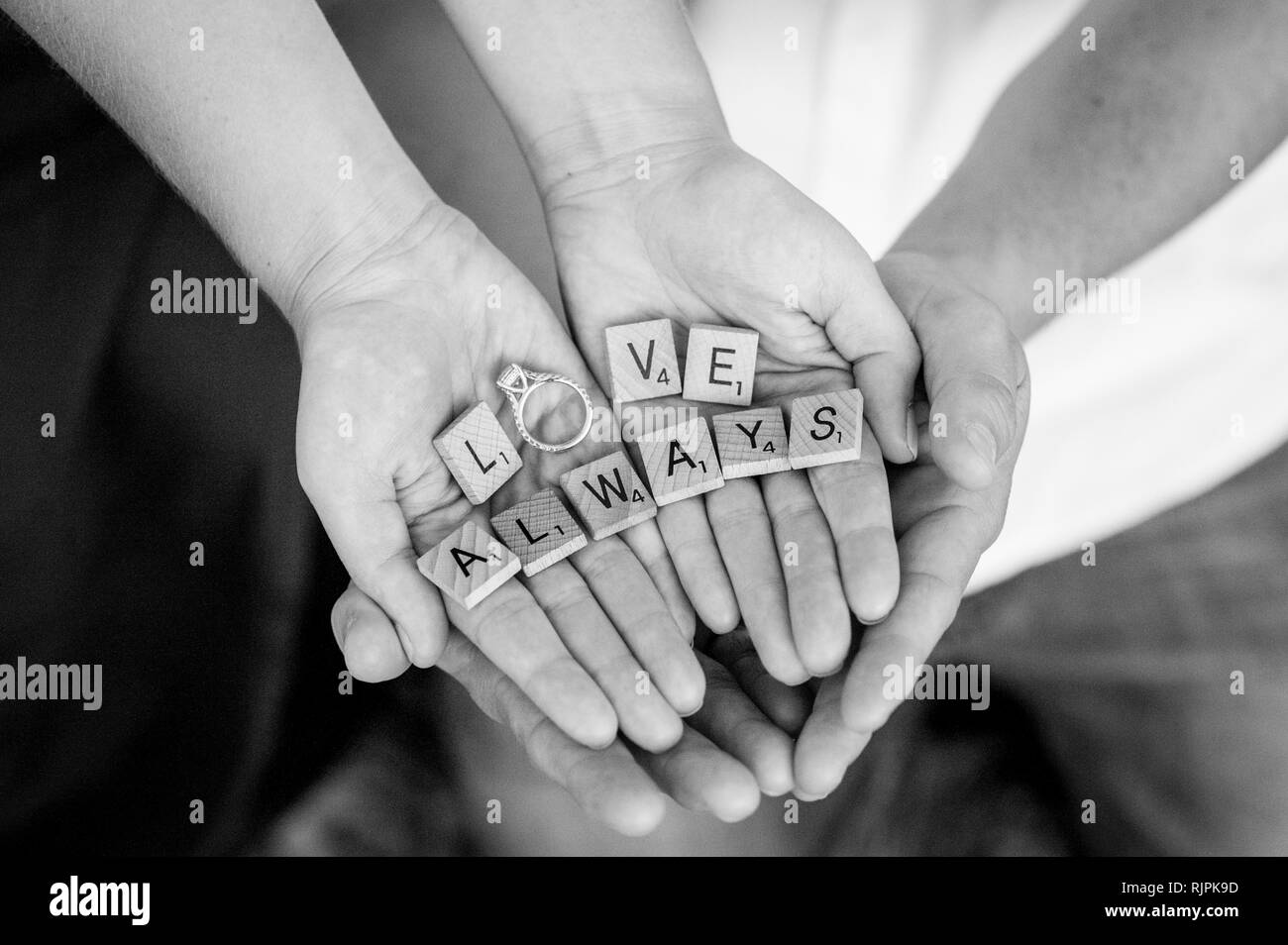 Horizontal close-up of an engaged couple's hands cupped around each other's, holding scrabble pieces spelling the words 'love always' with the ring. Stock Photo