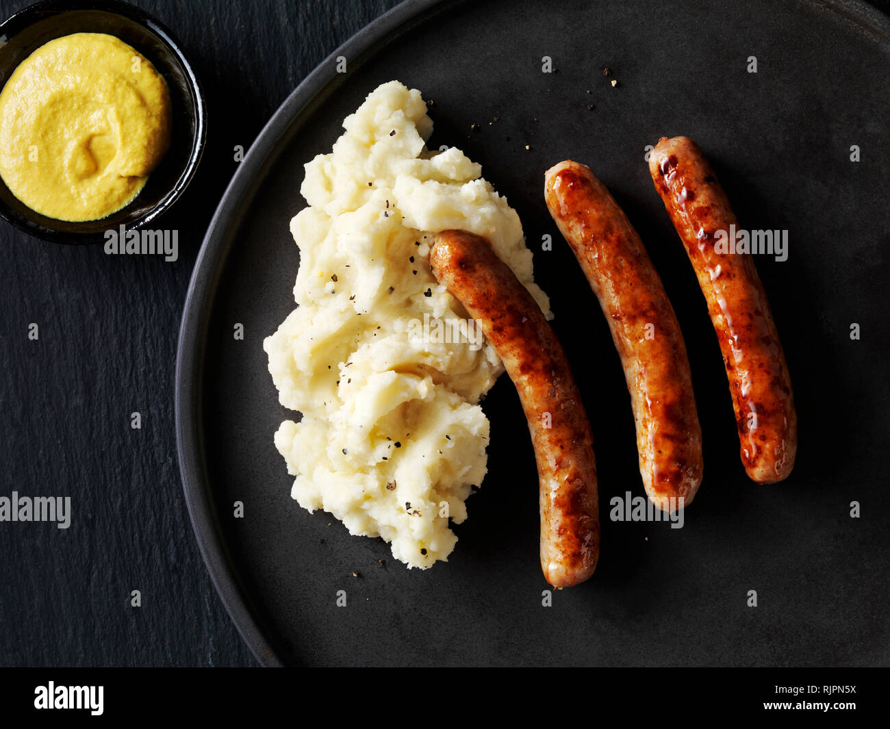 Still life with sausages and mash on black plate with bowl of mustard