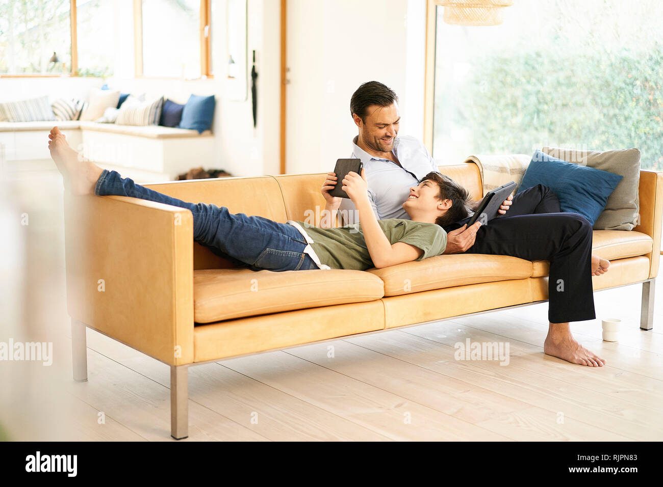Boy and father using digital tablets whilst relaxing on sofa Stock Photo