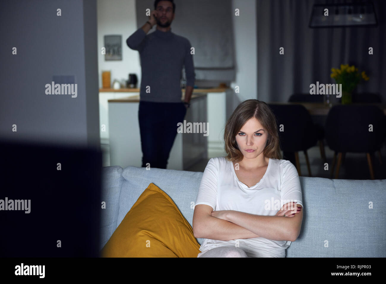 Young woman sulking on sofa in evening, while boyfriend makes smartphone call Stock Photo