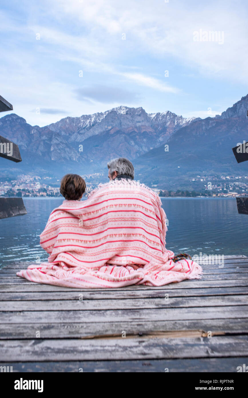 Boy and father wrapped in blanket on lakeside pier, rear view, Lake Como, Onno, Lombardy, Italy Stock Photo