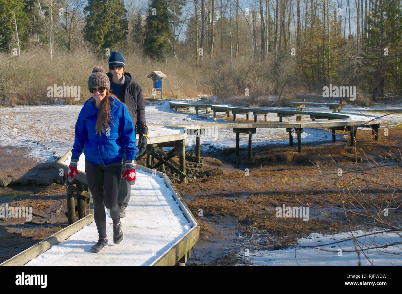 A smiling young couple walking the snowy boardwalk edging the mudflats at Shoreline Park, Port Moody, British Columbia, Canada. Stock Photo