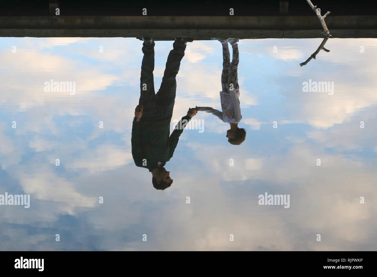 Reflection of father and daughter holding hands on a dock in the water amidst beautiful clouds Stock Photo