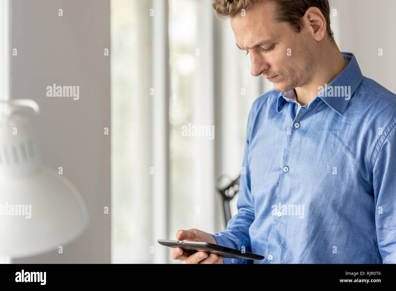 Serious young businessman standing by the office windows looking at his digital tablet. Stock Photo