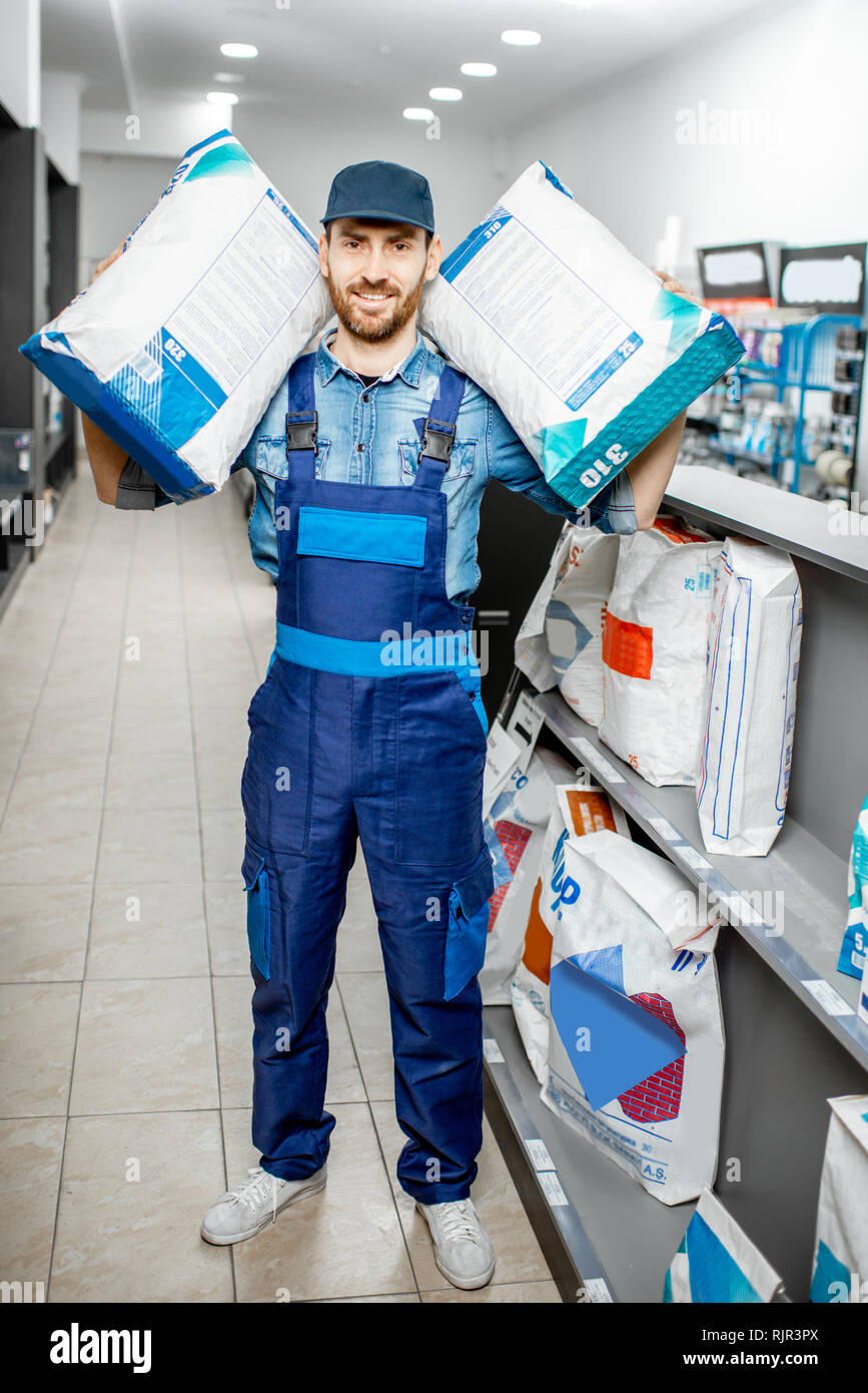 Portrait of a handsome workman in blue overalls holding bags with construction mixture in the building supermarket Stock Photo