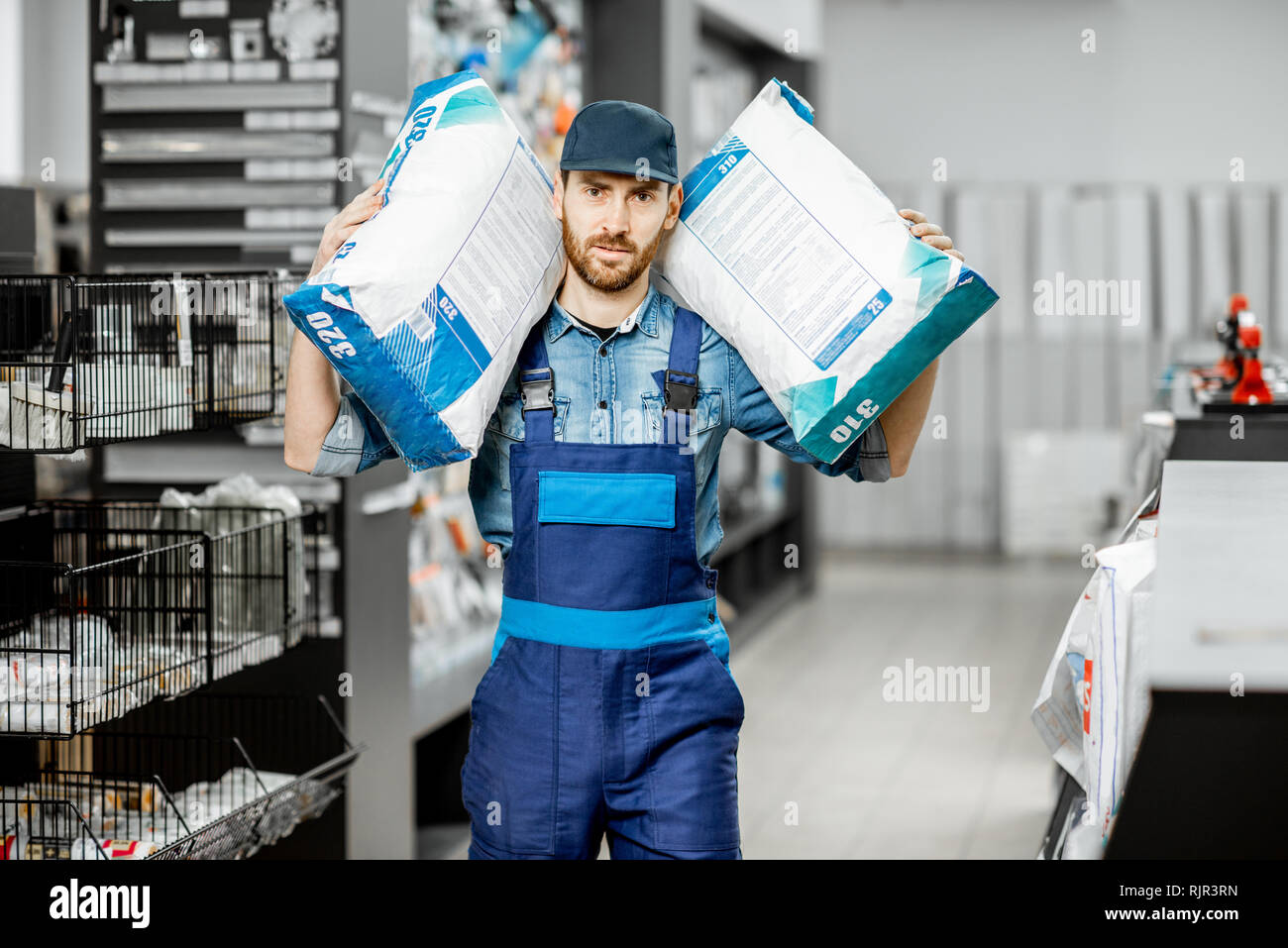 Portrait of a handsome workman in blue overalls holding bags with construction mixture in the building supermarket Stock Photo