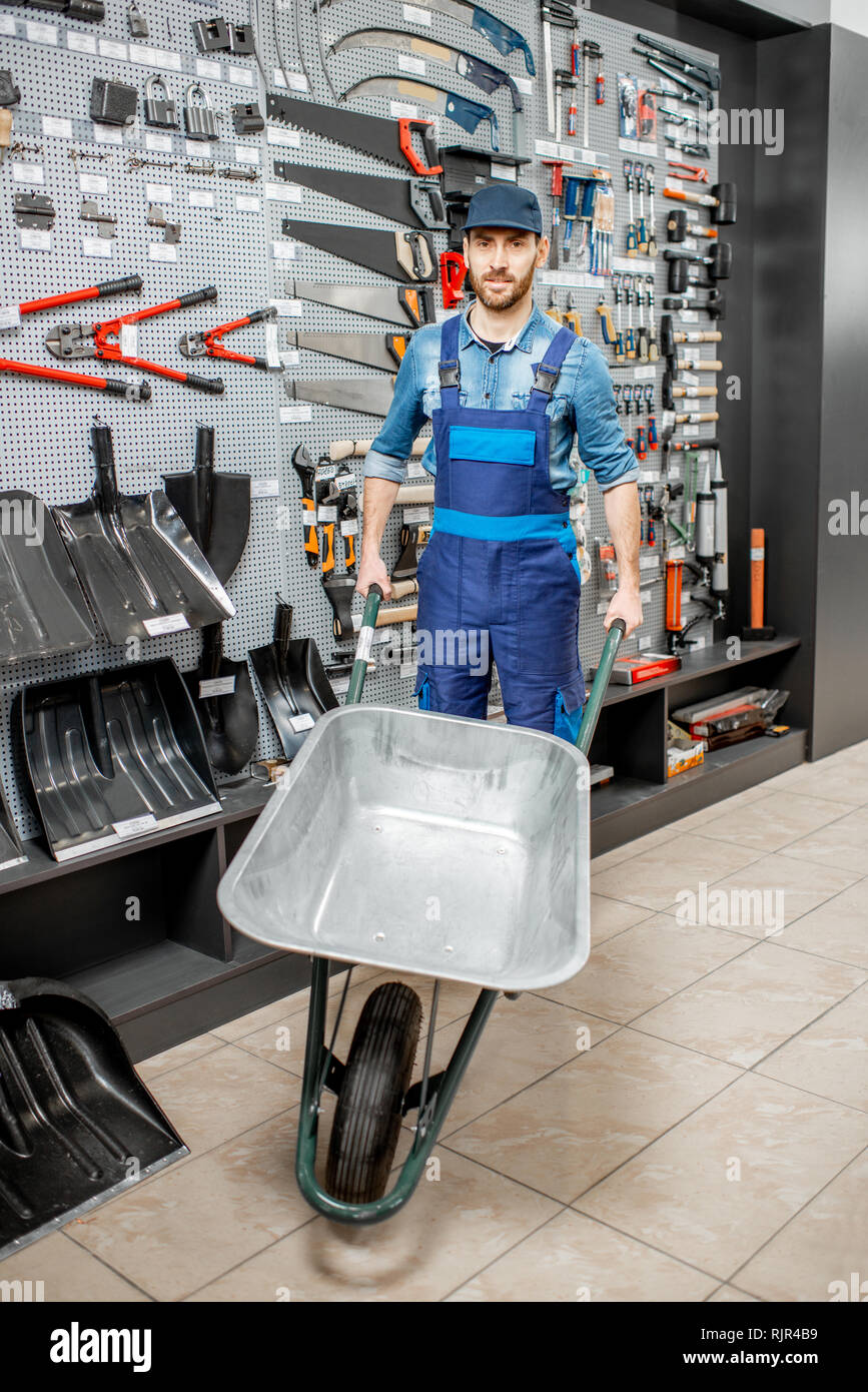 Portrait of a handsome worker in uniform standing with pushcart near the showcase with garden equipment in the shop Stock Photo