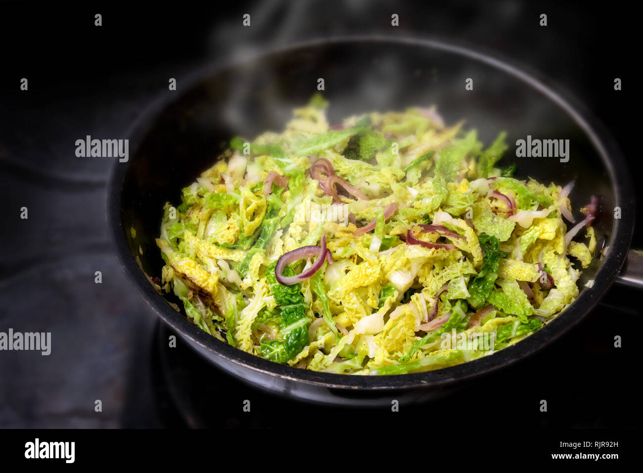 cooking green savoy cabbage with red onions in a black pan on a stove, healthy winter vegetable, selected focus, narrow depth of field Stock Photo