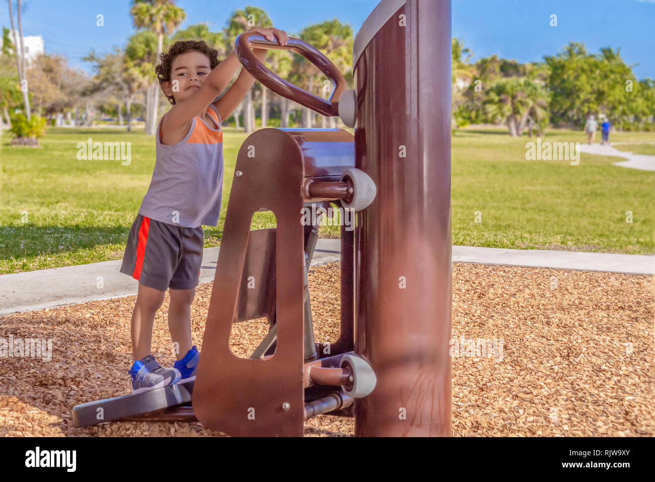 The young boy gets on the outdoor Stairmaster while looking over the other side for his mother.  The little boy keeps a watchful eye on mommy. Stock Photo