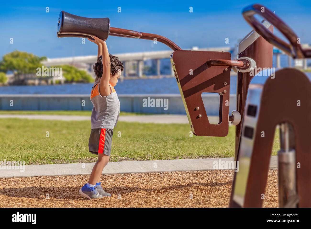 The little boy stretches high to reach the adult shoulder pads at the waterfront outdoor gym. Stock Photo