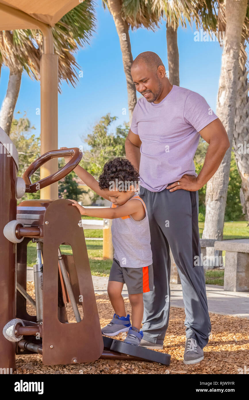 A father stands behind his little boy watching him use the Stairmaster at the outdoor gym. Stock Photo