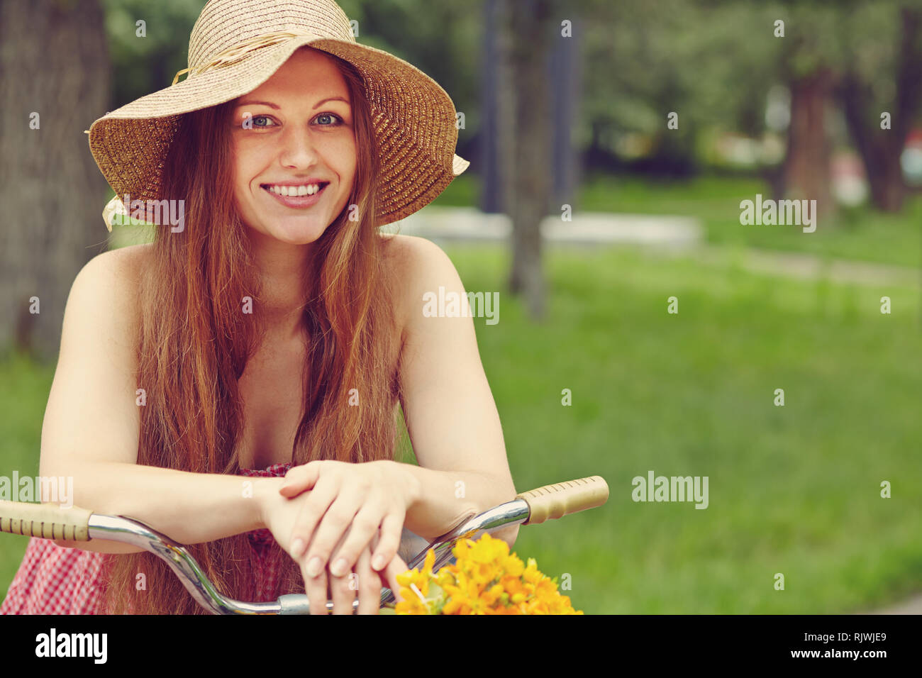 young woman in a dress and hat with a bike with flowers in a basket in a summer park Stock Photo