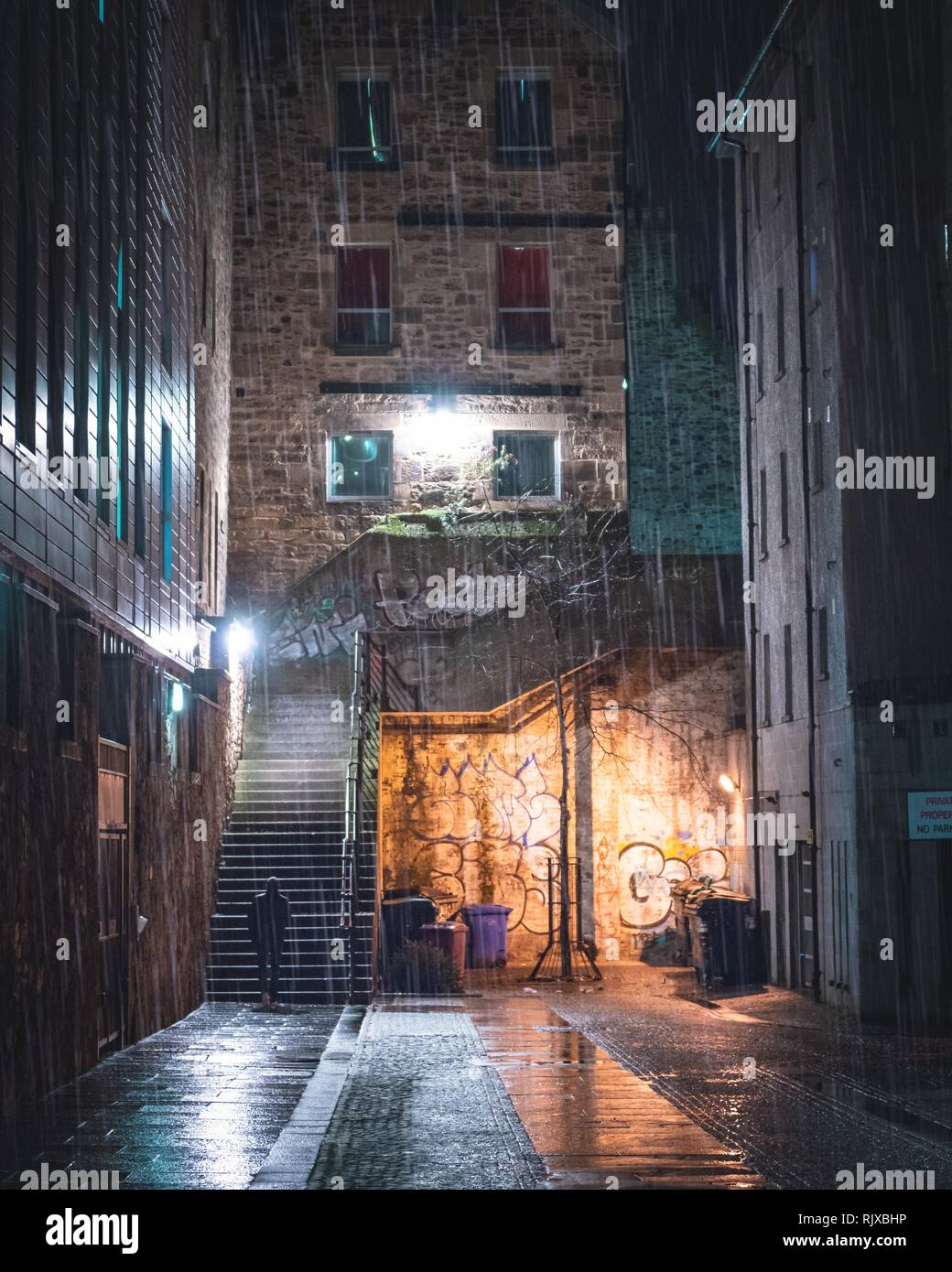 A singular figure about to climb some stairs down an alleyway in the city centre of Edinburgh, Scotland during the dark evening. The wet weather pours Stock Photo