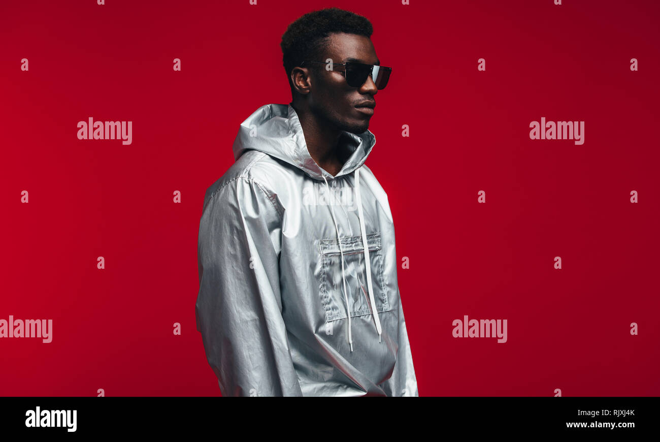 Stylish african man wearing shiny silver hooded shirt and sunglasses against red background. African american man in funky style outfit. Stock Photo